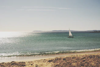 A serene Mediterranean coastline with a small sailboat gliding near rocky shores under a clear blue sky.