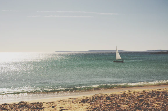 A serene Mediterranean coastline with a small sailboat gliding near rocky shores under a clear blue sky.