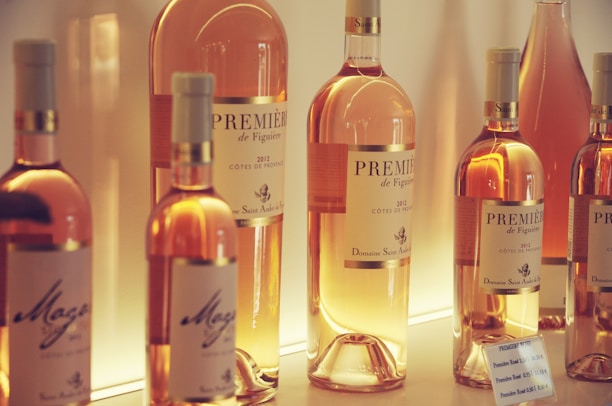 Several bottles of rosé wine are displayed on a well-lit shelf. The labels indicate they are from the Côtes de Provence region, with visible text such as 'Première de Figuière 2012' and 'Magali.' The bottles vary in size and are arranged in a visually appealing manner, showcasing their elegant design. The background lighting enhances the warm, inviting glow emanating from the wine.