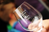 A hand holding a clear wine glass with white text printed on it. The text includes 'Domaine de l’onde' and 'Vins de Provence' accompanied by a small graphic. The background is blurred with hints of blue and purple, suggesting other people or objects out of focus.