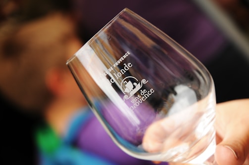 A hand holding a clear wine glass with white text printed on it. The text includes 'Domaine de l’onde' and 'Vins de Provence' accompanied by a small graphic. The background is blurred with hints of blue and purple, suggesting other people or objects out of focus.