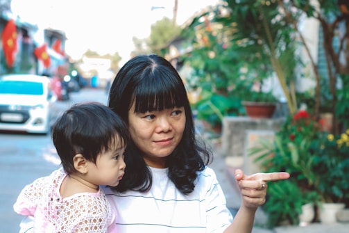 A woman holding a young child in her arms stands outdoors on a street lined with colorful flags and greenery. The woman is pointing at something with a gentle smile, while the child looks on curiously. The background features a parked car, plants, and buildings, giving a sense of an urban neighborhood.