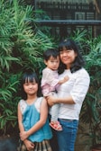 A woman holding a young child stands next to an older child. They are surrounded by lush green plants and stand in front of a window. The background features a mix of greenery and building architecture.