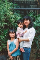 A woman holding a young child stands next to an older child. They are surrounded by lush green plants and stand in front of a window. The background features a mix of greenery and building architecture.