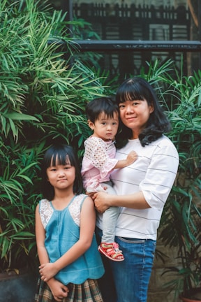 A woman holding a young child stands next to an older child. They are surrounded by lush green plants and stand in front of a window. The background features a mix of greenery and building architecture.