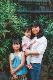 A woman holding a young child stands next to an older child. They are surrounded by lush green plants and stand in front of a window. The background features a mix of greenery and building architecture.