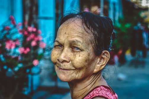 An elderly woman with an expressive face appears content and at peace. She is outdoors, with a blurred background of vibrant blue and pink tones, possibly flowers or a building.