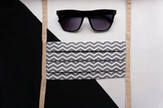 A pair of black sunglasses with dark lenses is placed above a striped fabric mask featuring a gray and white zigzag pattern. The background consists of diagonal black and white segments with beige straps bordering the mask.