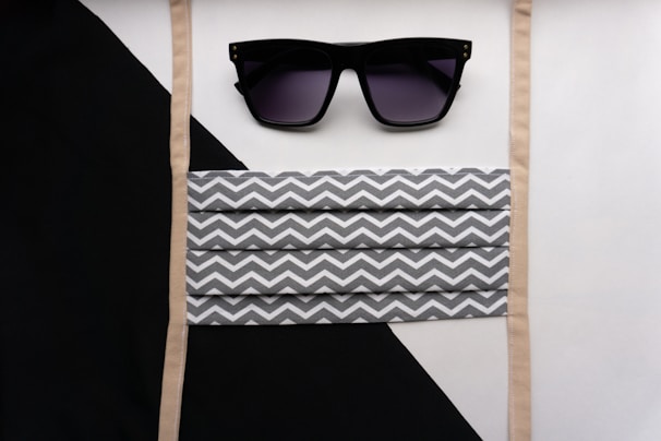 A pair of black sunglasses with dark lenses is placed above a striped fabric mask featuring a gray and white zigzag pattern. The background consists of diagonal black and white segments with beige straps bordering the mask.