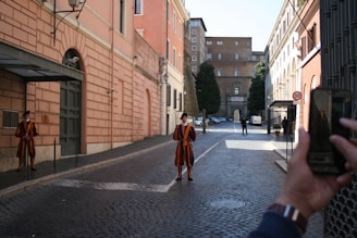 The image depicts a cobblestone street lined with historical buildings in muted shades of orange and cream. Two guards in bright, colorful uniforms with bold stripes stand at attention near the middle of the street. One person is visible in the foreground, capturing the scene with a smartphone. In the background, trees and a stone archway contribute to the serene urban setting.