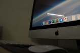 A close-up view of an Apple desktop computer displaying a screen with a digital workspace. The screen shows several application icons including browsers, productivity software, and messenger apps. The surrounding environment includes a keyboard and a mouse on a dark desk, suggesting a dimly lit room.