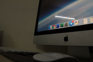 A close-up view of an Apple desktop computer displaying a screen with a digital workspace. The screen shows several application icons including browsers, productivity software, and messenger apps. The surrounding environment includes a keyboard and a mouse on a dark desk, suggesting a dimly lit room.