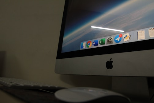 A close-up view of an Apple desktop computer displaying a screen with a digital workspace. The screen shows several application icons including browsers, productivity software, and messenger apps. The surrounding environment includes a keyboard and a mouse on a dark desk, suggesting a dimly lit room.