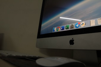 A close-up view of an Apple desktop computer displaying a screen with a digital workspace. The screen shows several application icons including browsers, productivity software, and messenger apps. The surrounding environment includes a keyboard and a mouse on a dark desk, suggesting a dimly lit room.