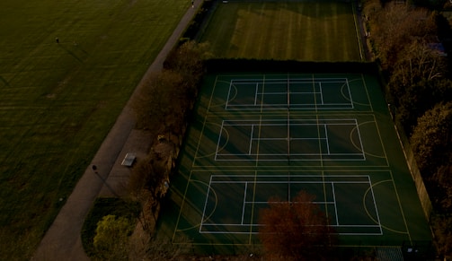 An aerial view of multiple tennis or sports courts next to a green recreational field, bordered by trees. The courts are marked with lines and look well-maintained. Shadows cast by trees stretch over parts of the courts and pathways.