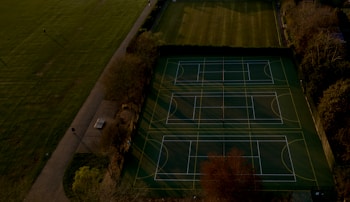 An aerial view of multiple tennis or sports courts next to a green recreational field, bordered by trees. The courts are marked with lines and look well-maintained. Shadows cast by trees stretch over parts of the courts and pathways.