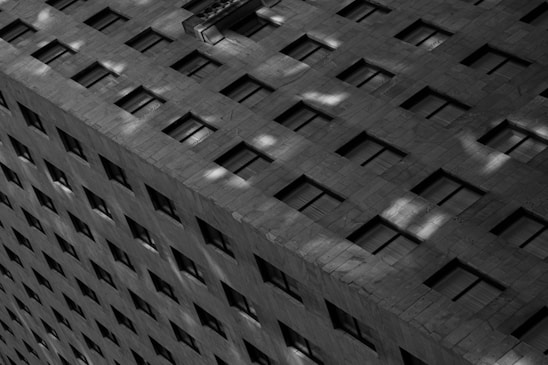 A monochromatic photograph of a high-rise building facade featuring a grid pattern of windows and concrete. The light casts sharp shadows and highlights, creating an abstract, geometric aesthetic.