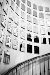 A series of small, angular mirrors arranged in a geometric pattern cover a white wall, reflecting various fragments of the surrounding space. In the lower part of the image, a curved staircase with dark iron railings leads gracefully downward, partially visible in the reflection.