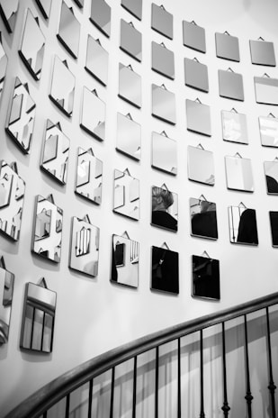 A series of small, angular mirrors arranged in a geometric pattern cover a white wall, reflecting various fragments of the surrounding space. In the lower part of the image, a curved staircase with dark iron railings leads gracefully downward, partially visible in the reflection.