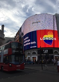 A vibrant urban scene with a large digital billboard displaying a pride-themed message and rainbow heart logo. A red double-decker bus is in the foreground, traveling on the city street. Several pedestrians can be observed walking nearby, and shops line the street.