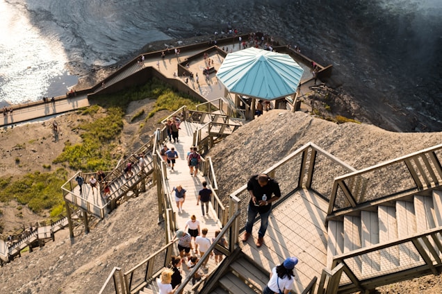 A steep wooden staircase with multiple platforms winds down a rocky hillside. Many people are walking up and down the steps, suggesting a popular tourist or scenic spot. At one platform, there's a structure with a teal roof providing some shelter or rest area. The area is partially surrounded by greenery and a body of water with reflections from the sun.