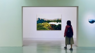 A person stands in an art gallery, facing a large landscape painting mounted on a white wall. The painting depicts a scenic view with green hills and blue mountains. It is surrounded by a minimalist gallery space with another smaller round artwork visible on the side wall.