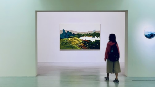 A person stands in an art gallery, facing a large landscape painting mounted on a white wall. The painting depicts a scenic view with green hills and blue mountains. It is surrounded by a minimalist gallery space with another smaller round artwork visible on the side wall.