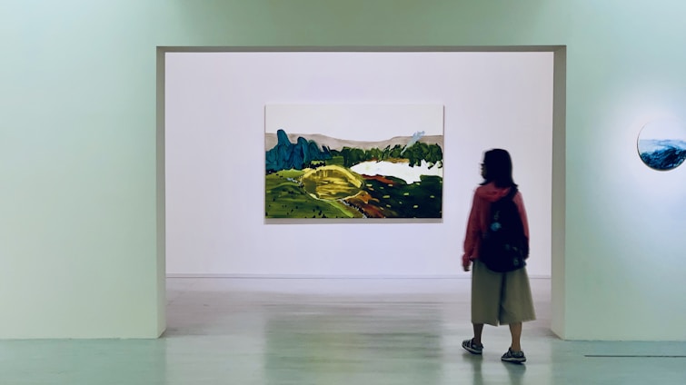 A person stands in an art gallery, facing a large landscape painting mounted on a white wall. The painting depicts a scenic view with green hills and blue mountains. It is surrounded by a minimalist gallery space with another smaller round artwork visible on the side wall.