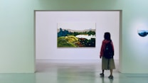 A person stands in an art gallery, facing a large landscape painting mounted on a white wall. The painting depicts a scenic view with green hills and blue mountains. It is surrounded by a minimalist gallery space with another smaller round artwork visible on the side wall.