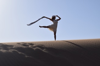 A person wearing a dress performs a graceful dance or yoga pose on a sand dune, holding a flowing scarf in one hand. The scene is captured at dawn or dusk, creating a dramatic silhouette against a clear sky.