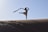 A person wearing a dress performs a graceful dance or yoga pose on a sand dune, holding a flowing scarf in one hand. The scene is captured at dawn or dusk, creating a dramatic silhouette against a clear sky.