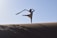 A person wearing a dress performs a graceful dance or yoga pose on a sand dune, holding a flowing scarf in one hand. The scene is captured at dawn or dusk, creating a dramatic silhouette against a clear sky.