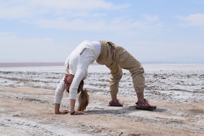 A person dressed in a white long-sleeved top and beige pants is performing a backbend on a dry, cracked surface, possibly a salt flat. The footwear is sturdy, suitable for outdoor terrain. The sky above is partly cloudy and blue, creating a contrast with the light-colored ground.