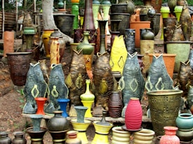 A vibrant collection of pottery and ceramic vases featuring fish-shaped designs and traditional shapes. The ceramics are arranged outdoors on a natural dirt surface with a background of trees. The vases exhibit various textures and colors, including earthy tones as well as vibrant greens, yellows, reds, and blues.