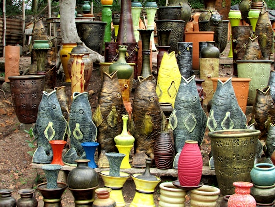 A vibrant collection of pottery and ceramic vases featuring fish-shaped designs and traditional shapes. The ceramics are arranged outdoors on a natural dirt surface with a background of trees. The vases exhibit various textures and colors, including earthy tones as well as vibrant greens, yellows, reds, and blues.