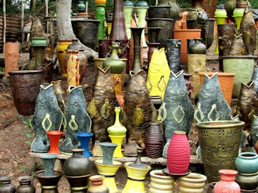 A vibrant collection of pottery and ceramic vases featuring fish-shaped designs and traditional shapes. The ceramics are arranged outdoors on a natural dirt surface with a background of trees. The vases exhibit various textures and colors, including earthy tones as well as vibrant greens, yellows, reds, and blues.