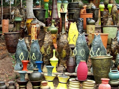 A vibrant collection of pottery and ceramic vases featuring fish-shaped designs and traditional shapes. The ceramics are arranged outdoors on a natural dirt surface with a background of trees. The vases exhibit various textures and colors, including earthy tones as well as vibrant greens, yellows, reds, and blues.