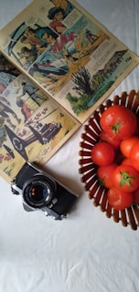 A vintage comic book is open on a white surface, showcasing colorful illustrations and speech bubbles. Next to the comic is an old-fashioned camera with a lens cap displaying text. A wicker basket filled with ripe red tomatoes sits nearby, adding a pop of color to the scene.
