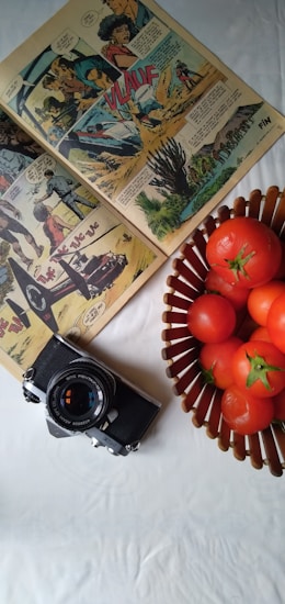 A vintage comic book is open on a white surface, showcasing colorful illustrations and speech bubbles. Next to the comic is an old-fashioned camera with a lens cap displaying text. A wicker basket filled with ripe red tomatoes sits nearby, adding a pop of color to the scene.