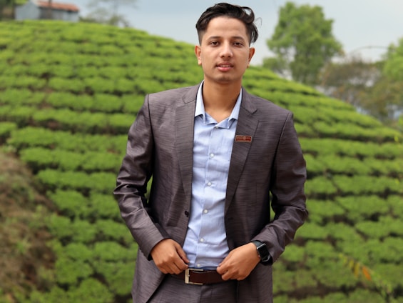 A person wearing a gray suit jacket and light blue shirt stands in front of a lush, green, terraced landscape. There is a small building in the background and trees are visible, suggesting a natural, outdoor setting.