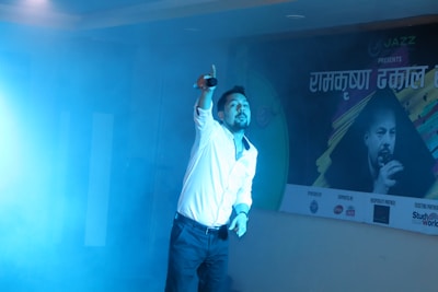A person holding a microphone appears to be performing on stage amidst a blue mist. The individual is wearing a white shirt and dark pants, with one arm stretched upwards. Behind them, a banner displays text in a non-English script alongside an image of another person holding a microphone.