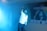 A person holding a microphone appears to be performing on stage amidst a blue mist. The individual is wearing a white shirt and dark pants, with one arm stretched upwards. Behind them, a banner displays text in a non-English script alongside an image of another person holding a microphone.