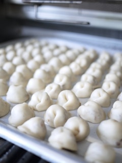 Rows of uncooked dumplings are arranged neatly on a baking tray, ready for cooking. The background shows a blurred view of a kitchen environment, suggesting a setting of food preparation.