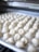 Rows of uncooked dumplings are arranged neatly on a baking tray, ready for cooking. The background shows a blurred view of a kitchen environment, suggesting a setting of food preparation.