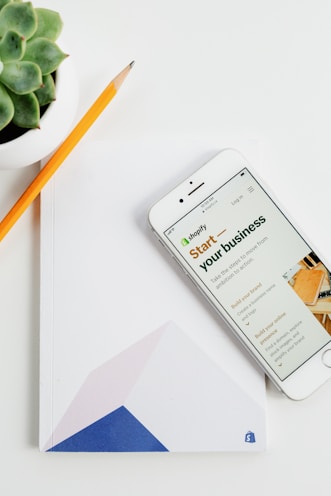 A smartphone displaying a Shopify webpage with the text 'Start your business' rests on a white surface. Next to it lies a closed notebook with geometric shapes on its cover and a sharpened pencil. A small succulent plant is visible in the top left corner.