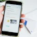A hand holds a smartphone displaying the Shopify website, focusing on a page about payment processing. Text about Simple Payments and Shopify Payments is visible along with some graphical elements like a chair and a desk lamp.