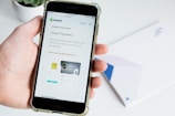 A hand holds a smartphone displaying the Shopify website, focusing on a page about payment processing. Text about Simple Payments and Shopify Payments is visible along with some graphical elements like a chair and a desk lamp.