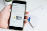 A hand holds a smartphone displaying the Shopify website, focusing on a page about payment processing. Text about Simple Payments and Shopify Payments is visible along with some graphical elements like a chair and a desk lamp.