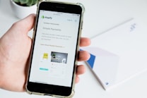 A hand holds a smartphone displaying the Shopify website, focusing on a page about payment processing. Text about Simple Payments and Shopify Payments is visible along with some graphical elements like a chair and a desk lamp.