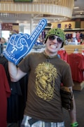 A person in a store wearing a brown graphic t-shirt, plaid shorts, sunglasses, and a knit hat. They are holding a large blue foam hand with 'Rangers' on it, and have a wide grin. The store appears to have sports merchandise, visible in the background.